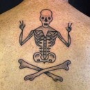 tattoo of small skeleton sitting cross legged giving the peace hand symbol